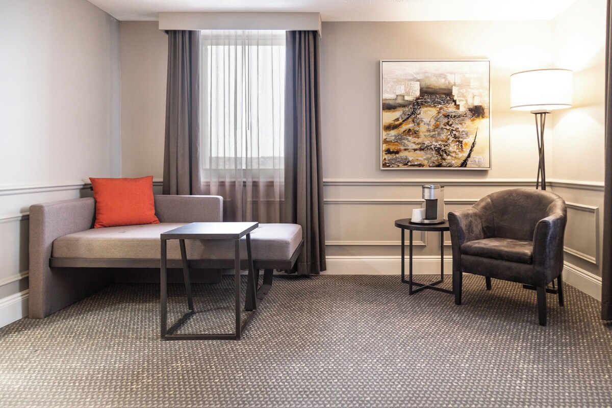 Een hotelkamer met een bank, fauteuil, klein tafeltje, lamp en kunst aan de muur.