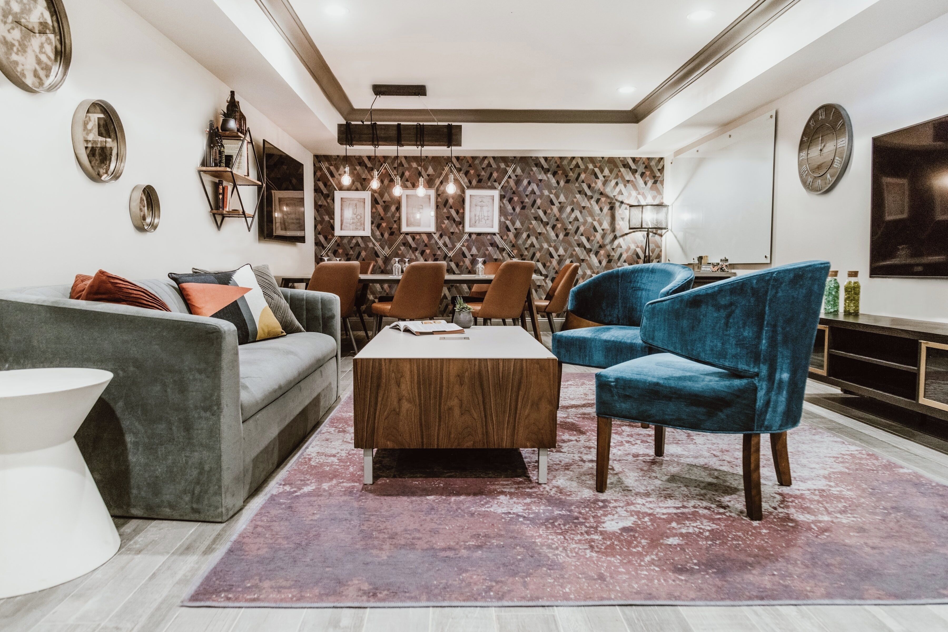 A modern living room with a grey sofa, a wooden coffee table, and blue velvet armchairs. There is a dining area with a wooden table and chairs, and a kitchen area with a TV and shelves.