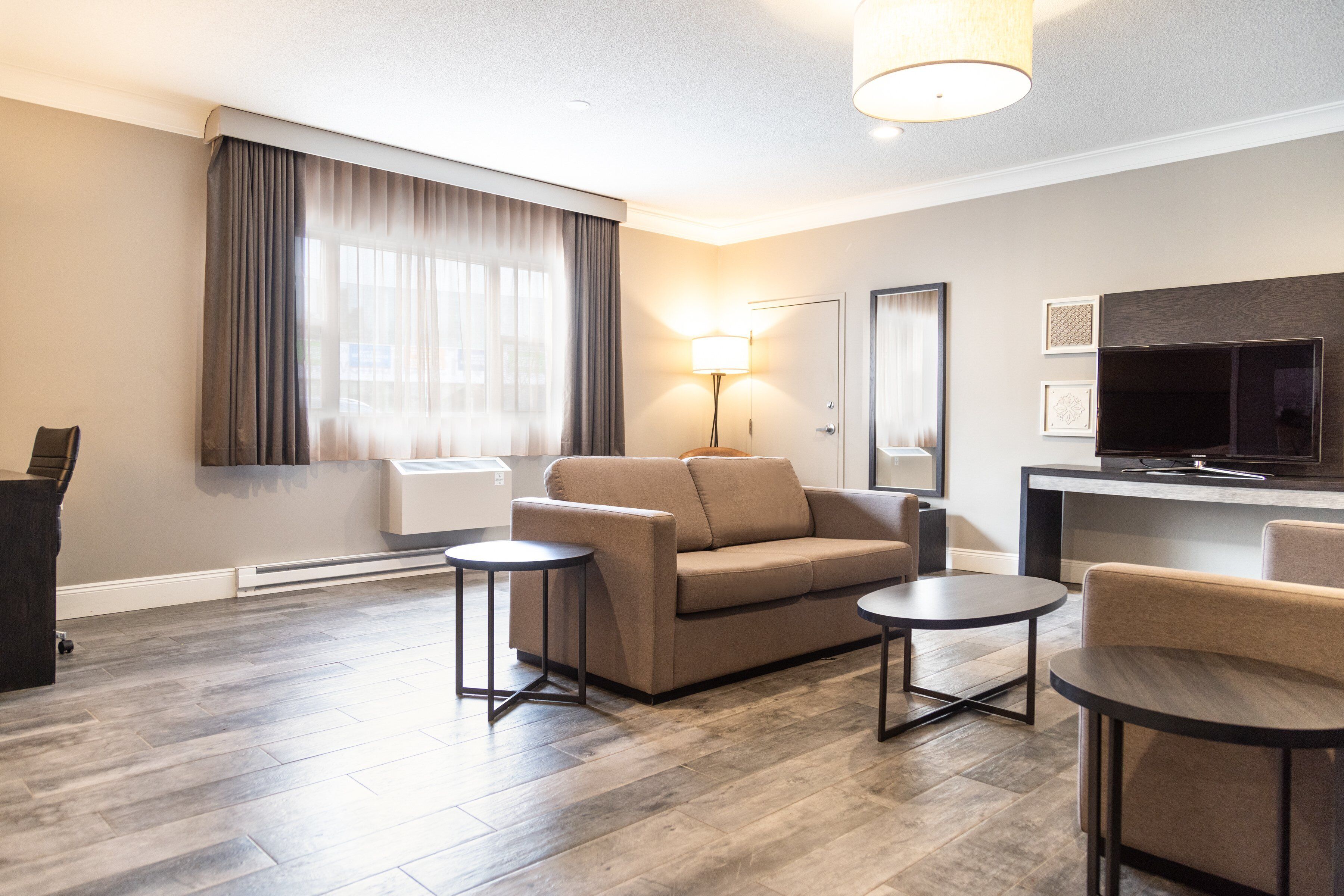 A modern hotel room with a sofa, two small tables, a flat-screen TV, and a lamp.