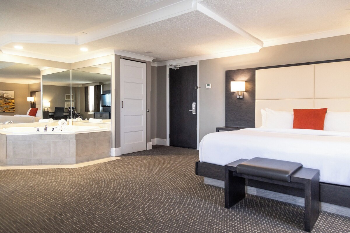 Een hotelkamer met een groot bed, een aparte badkamer met een badkuip, en een spiegel die de kamer weerspiegelt.