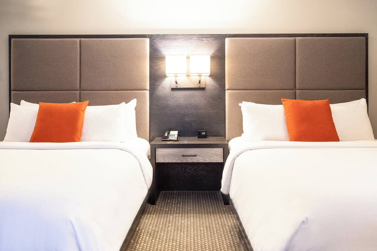 Une chambre d’hôtel avec deux lits, chacun doté de draps blancs et d’oreillers orange, une table de chevet avec un téléphone et une petite table, ainsi qu’un luminaire fixé au mur.
