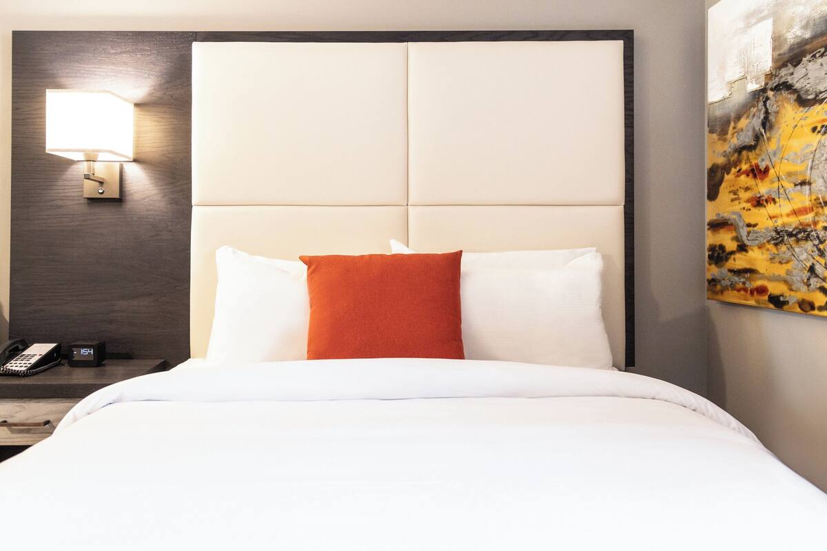 A hotel room with a large bed, a nightstand with a phone and clock, a wall-mounted lamp, and a painting on the wall.