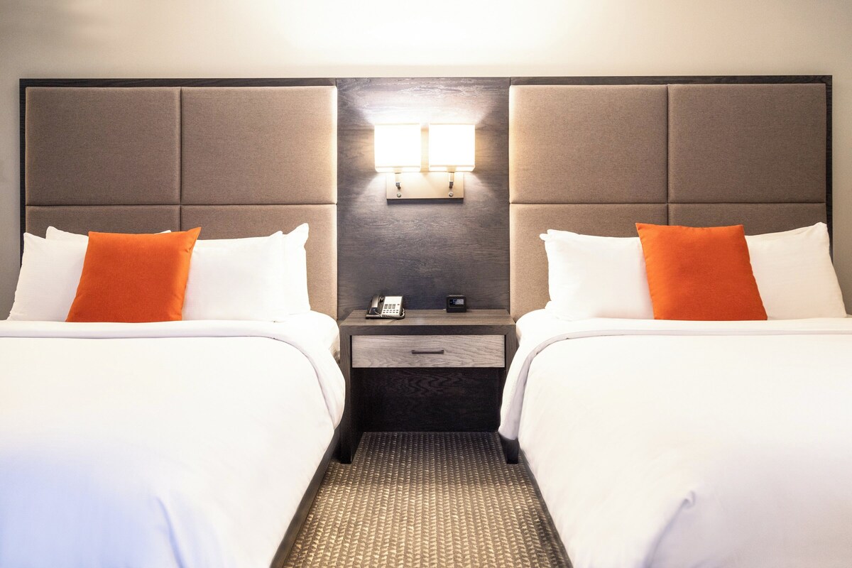Een hotelkamer met twee bedden, elk voorzien van witte lakens en oranje kussens, een nachtkastje met een telefoon en een klein tafeltje, en een wandlamp.