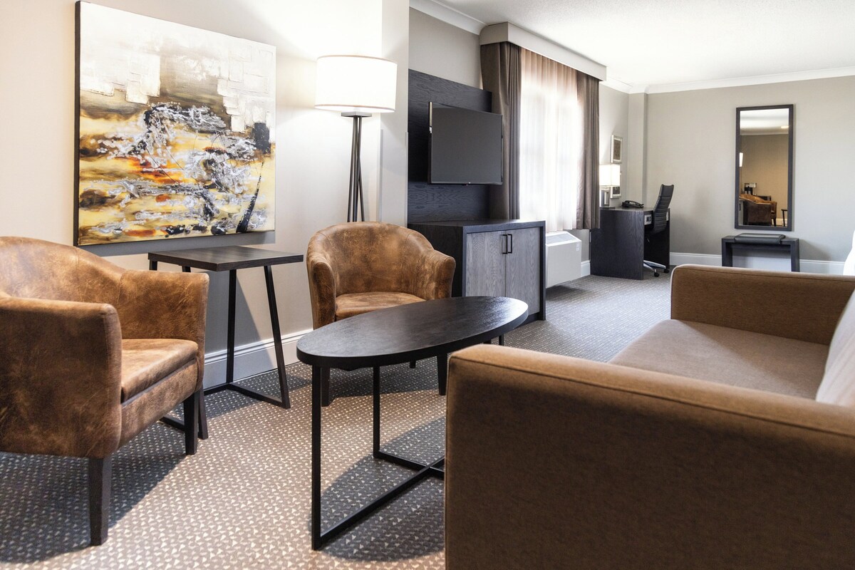 Een moderne hotelkamer met een sofa, twee fauteuils, een kleine tafel, een flatscreen televisie, een bureau en een groot abstract schilderij.