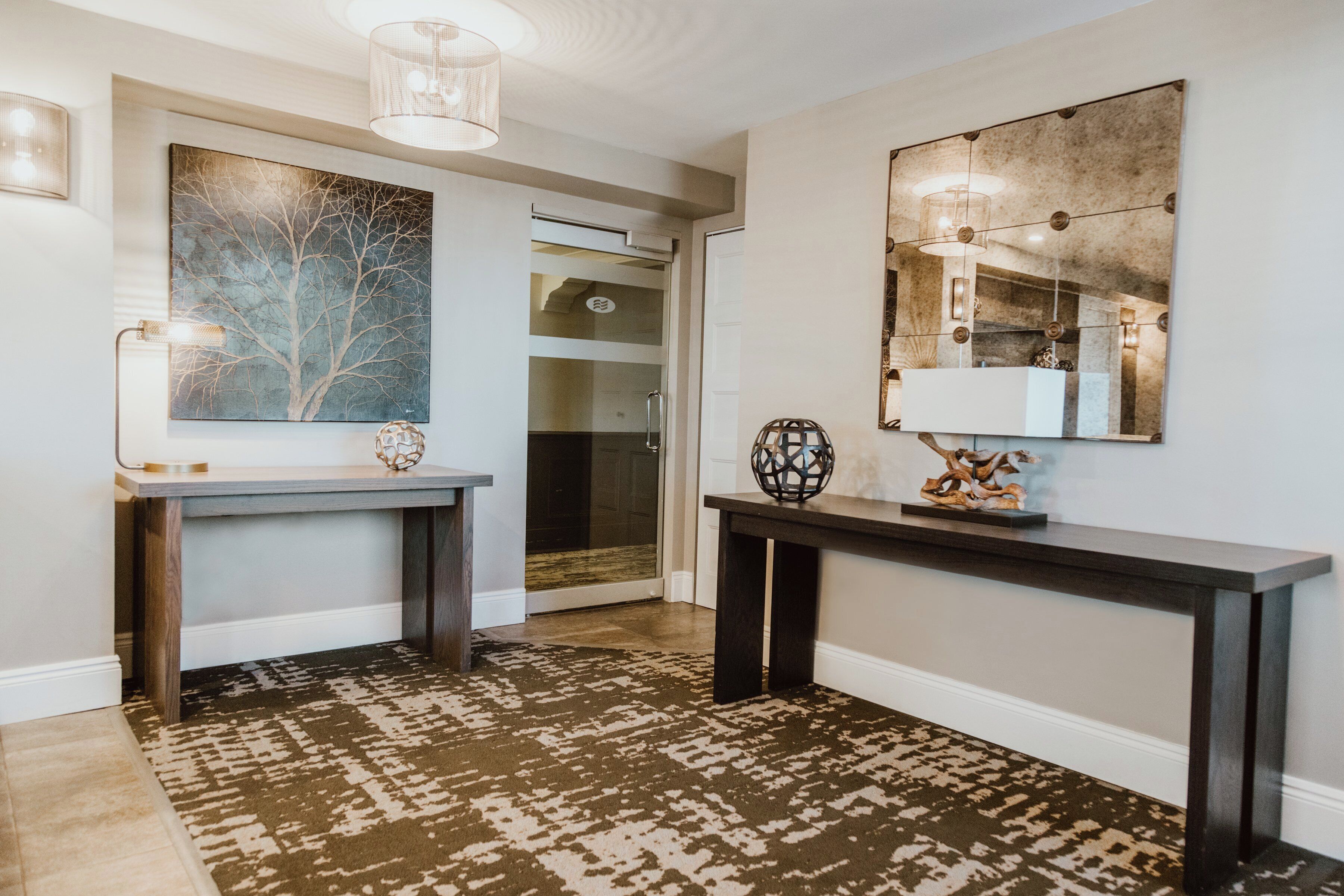 A modern hallway with a patterned rug, a console table with decorative items, a large abstract painting, and a mirror with lights.