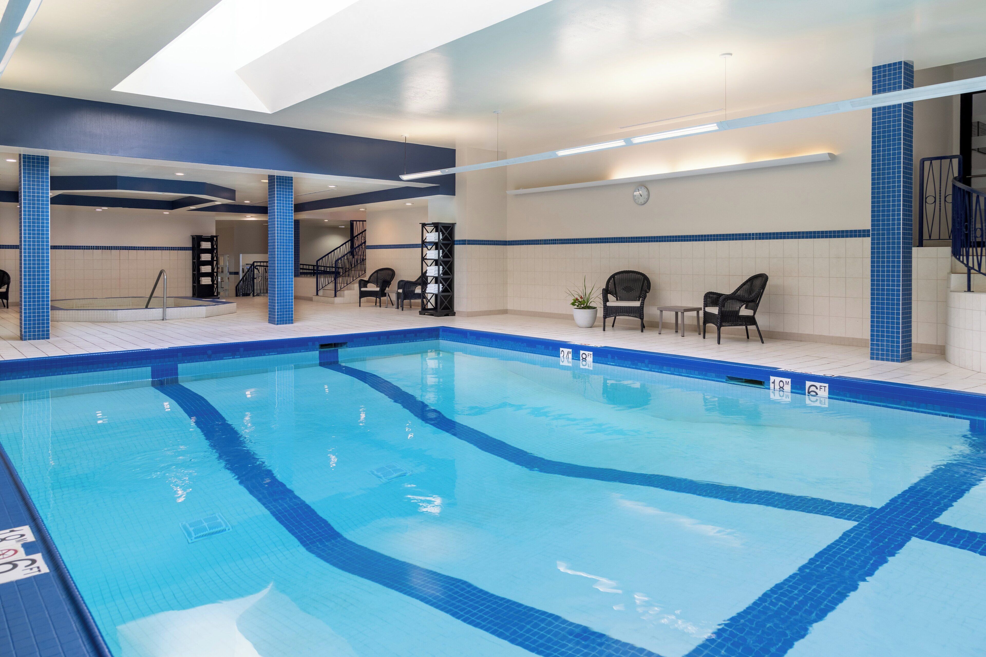 Piscina interior com faixas delimitadoras bem visíveis, piso em mosaico e área de estar com cadeiras e uma pequena mesa.