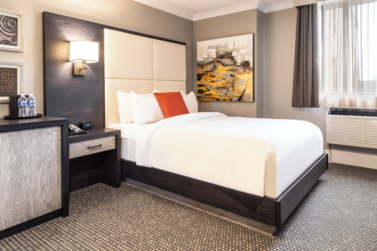 Een hotelkamer met een groot bed, een nachtkastje met een lamp, een bureau met een stoel, en een schilderij aan de muur.