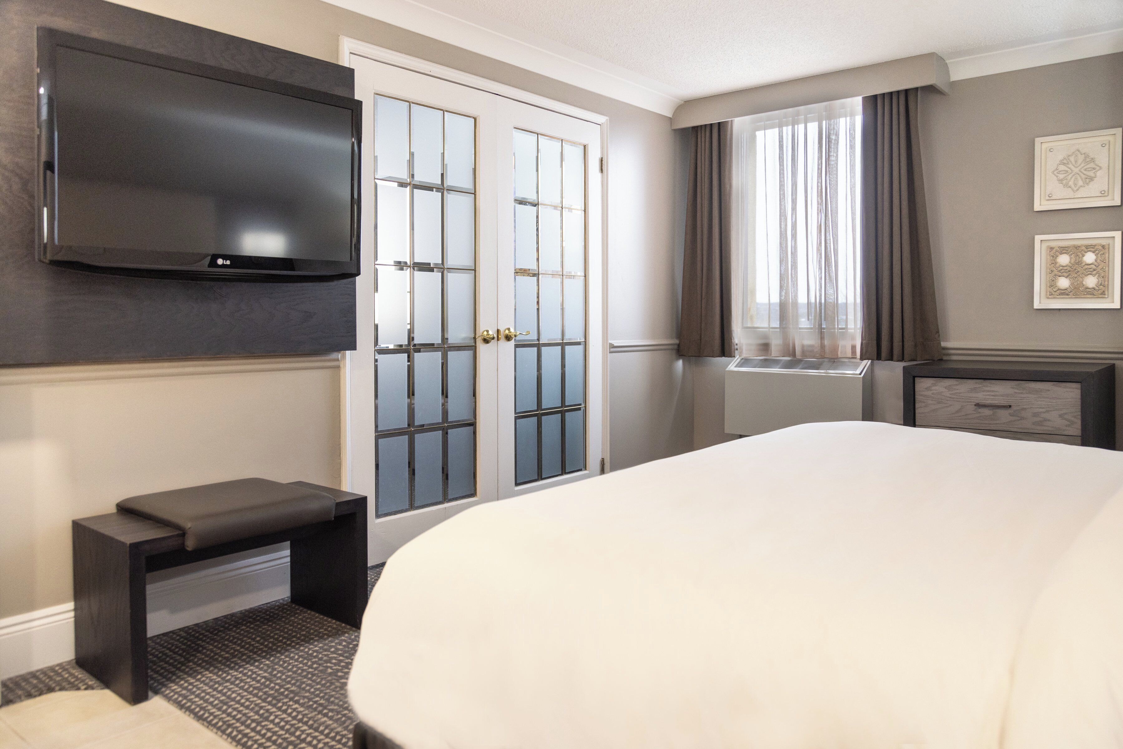 A hotel room with a large bed, a flat-screen TV mounted on the wall, a bench, a window with curtains, and a dresser.