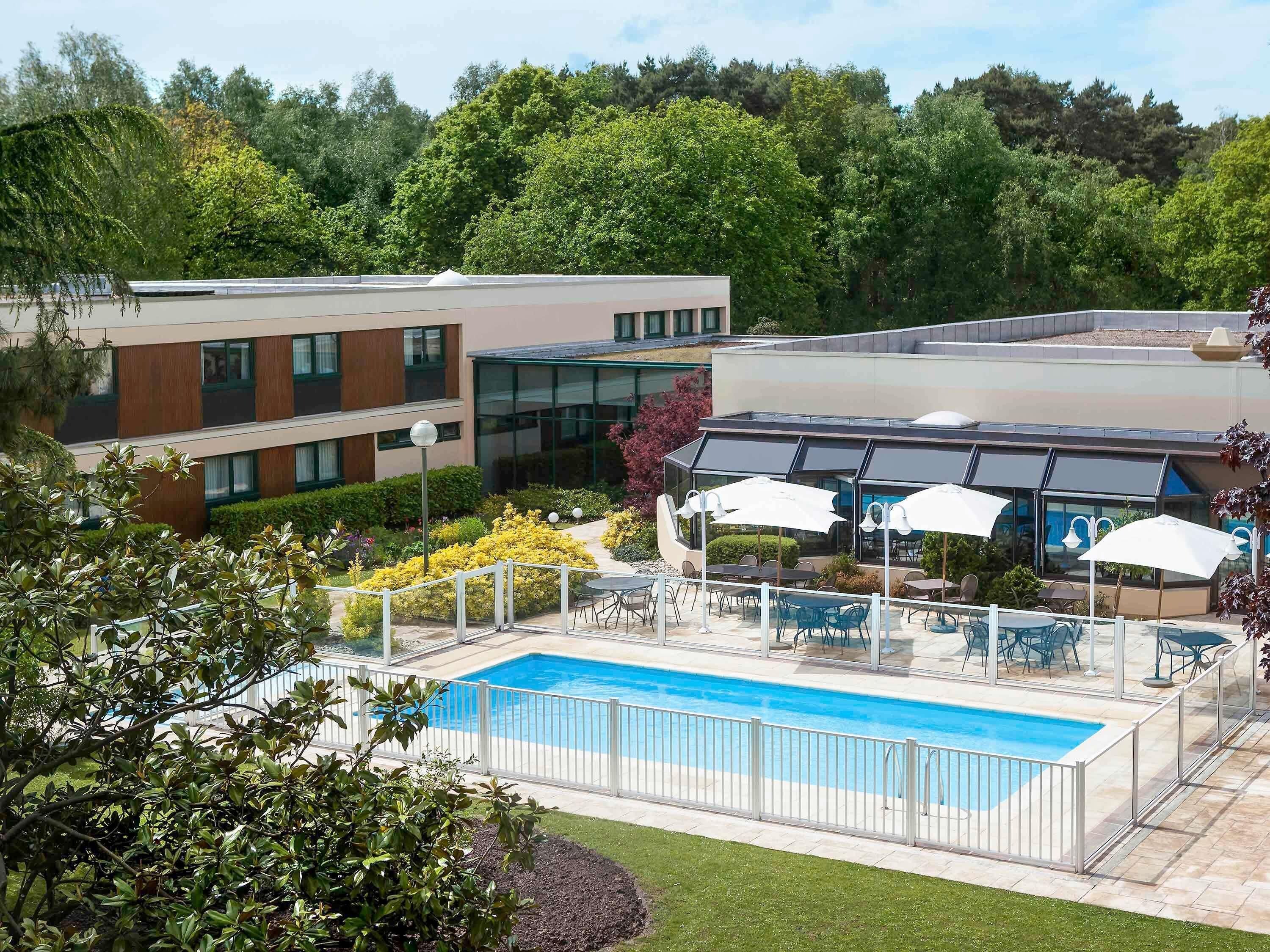 A swimming pool with a white fence, outdoor seating with umbrellas, and a building with multiple windows and a glass facade.