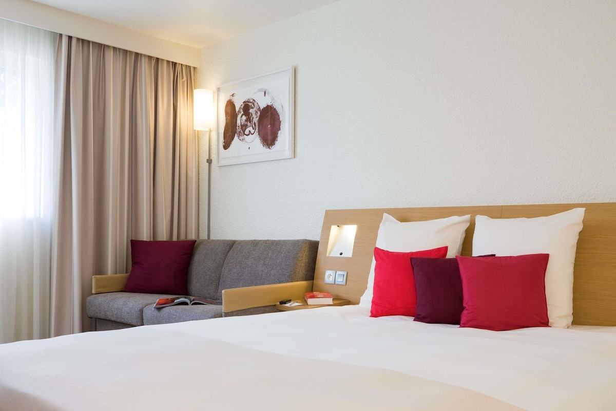 A hotel room with a large bed, a sofa, a bedside table, and a wall-mounted artwork.