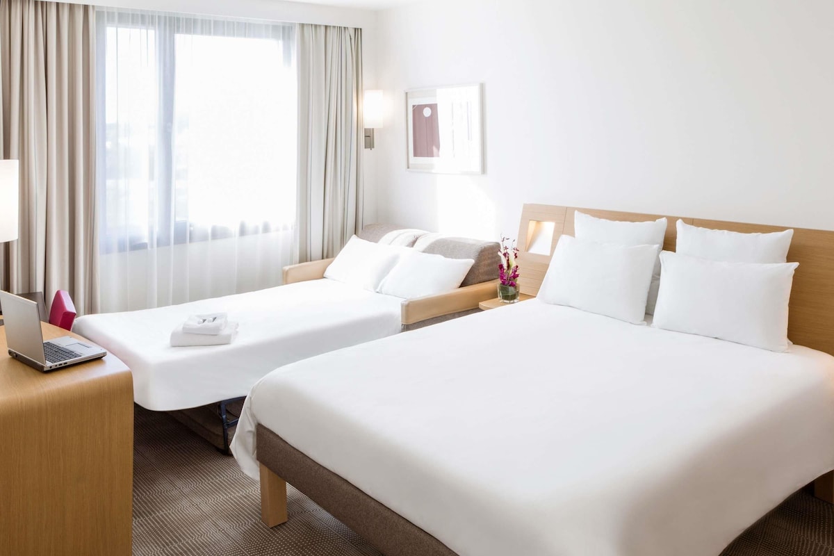 A hotel room with two beds, a desk with a laptop, a nightstand, and a window with curtains.