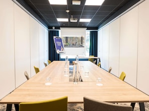Un salón de conferencias con una mesa larga, sillas, una pantalla de proyector y un ventanal con cortinas.