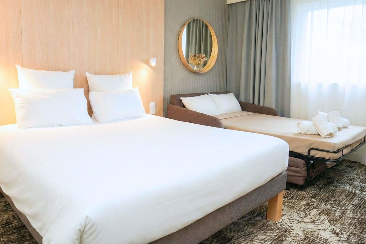 A hotel room with a large bed, a smaller bed, a sofa bed, a round mirror, and a window with curtains.