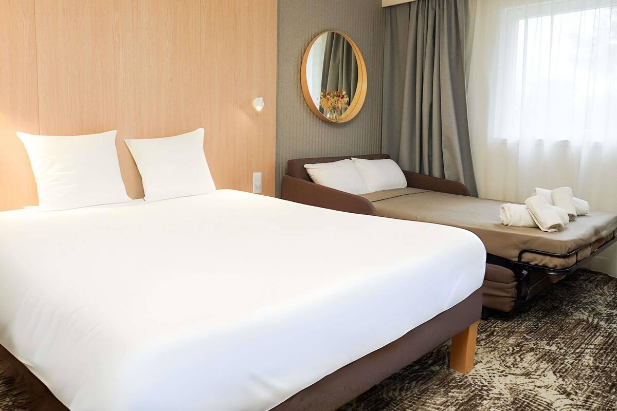 Una habitación de hotel con una cama grande, un sofá cama más pequeño, un espejo y una ventana con cortinas.