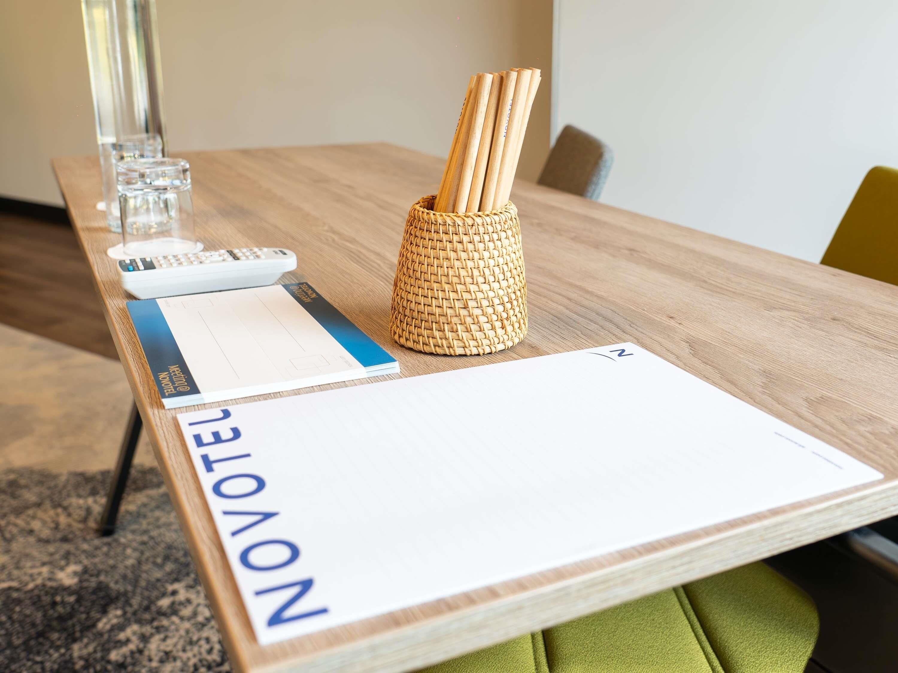 A hotel room with a wooden table, a woven basket with wooden sticks, a glass, a calculator, and a check-in form.