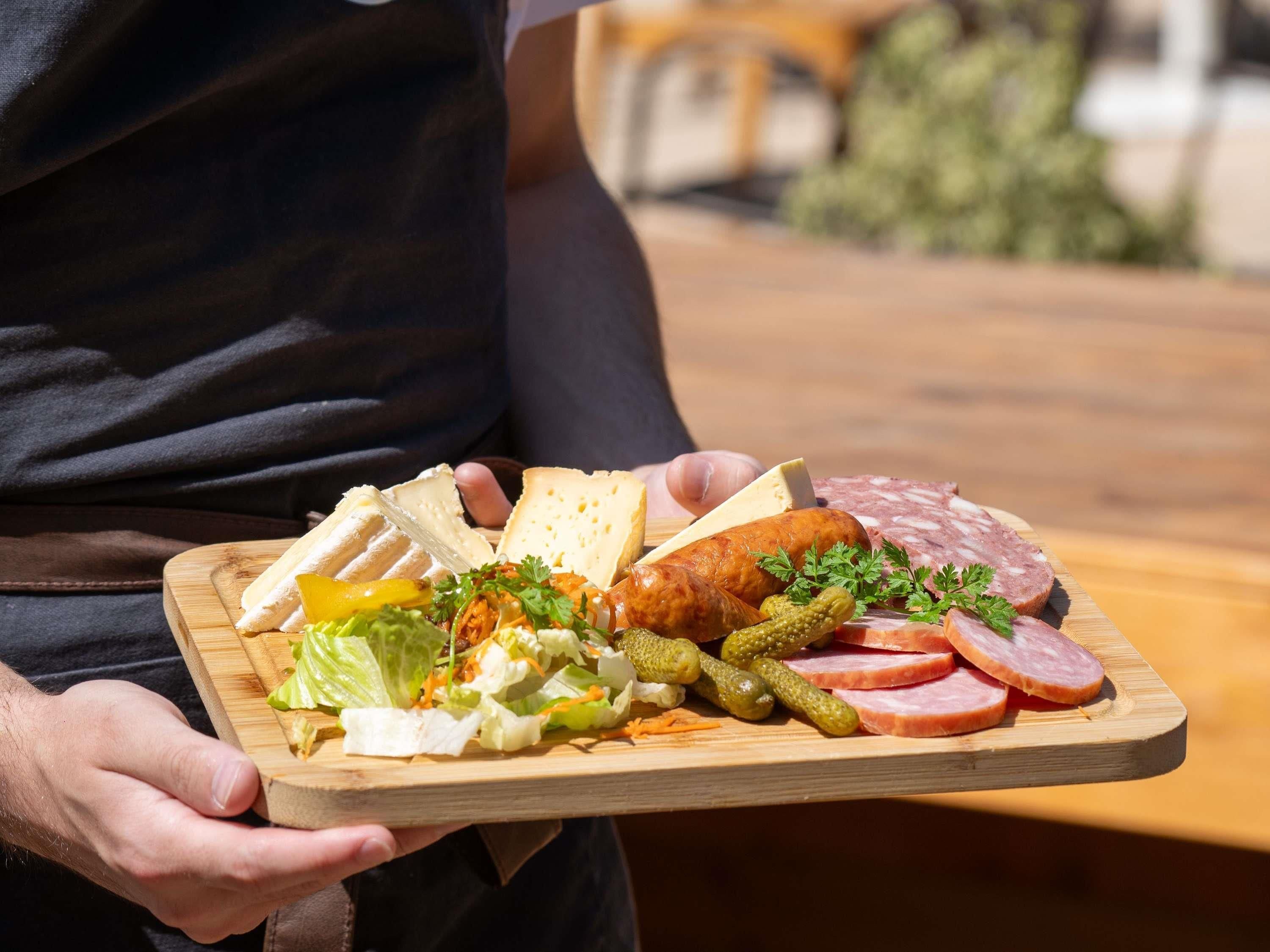 Una tabla de madera con diversos alimentos como pan, queso y embutidos, sostenida por una persona.