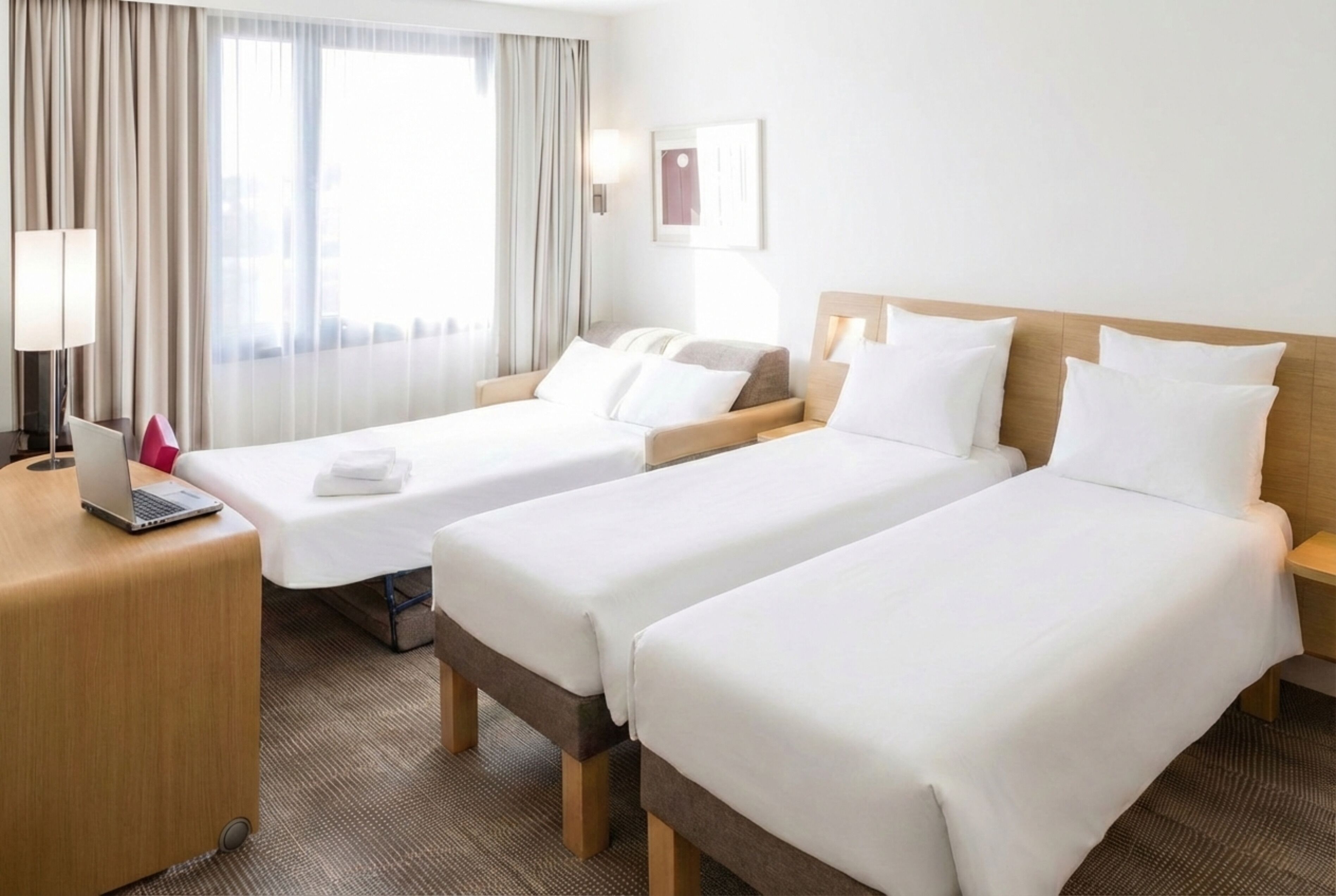A hotel room with two single beds, a desk with a laptop, and a large window with curtains.