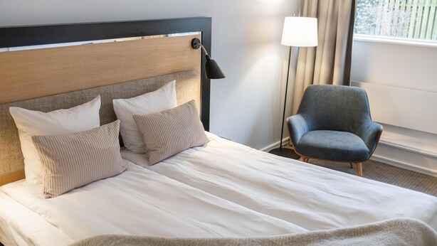 A neatly made bed with pillows, a bedside lamp, a chair, and a window with curtains. - Gentofte Hotel (Gentofte)