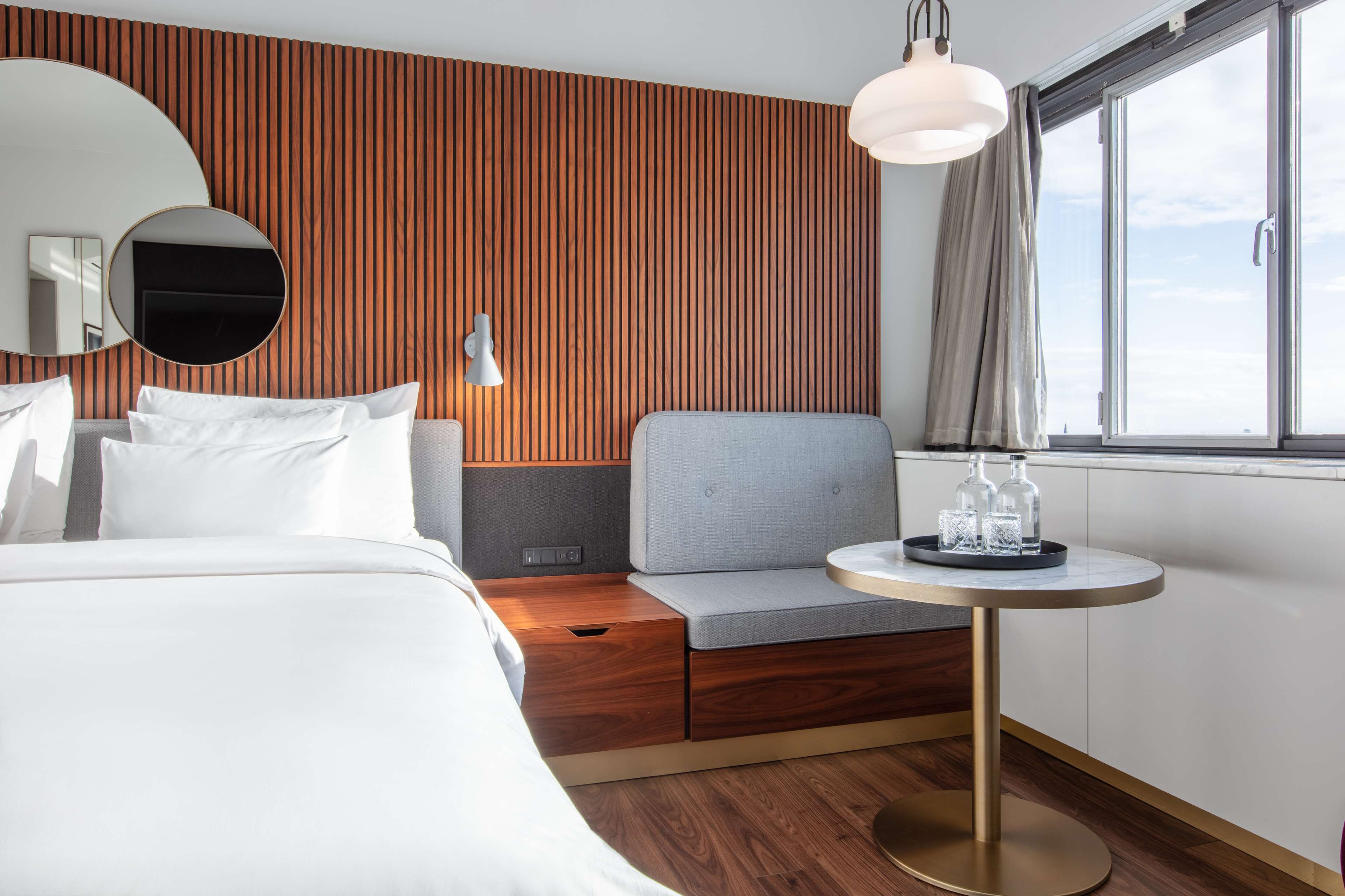 Una habitación de hotel moderna con cama, zona de estar, una mesa redonda y un ventanal grande.