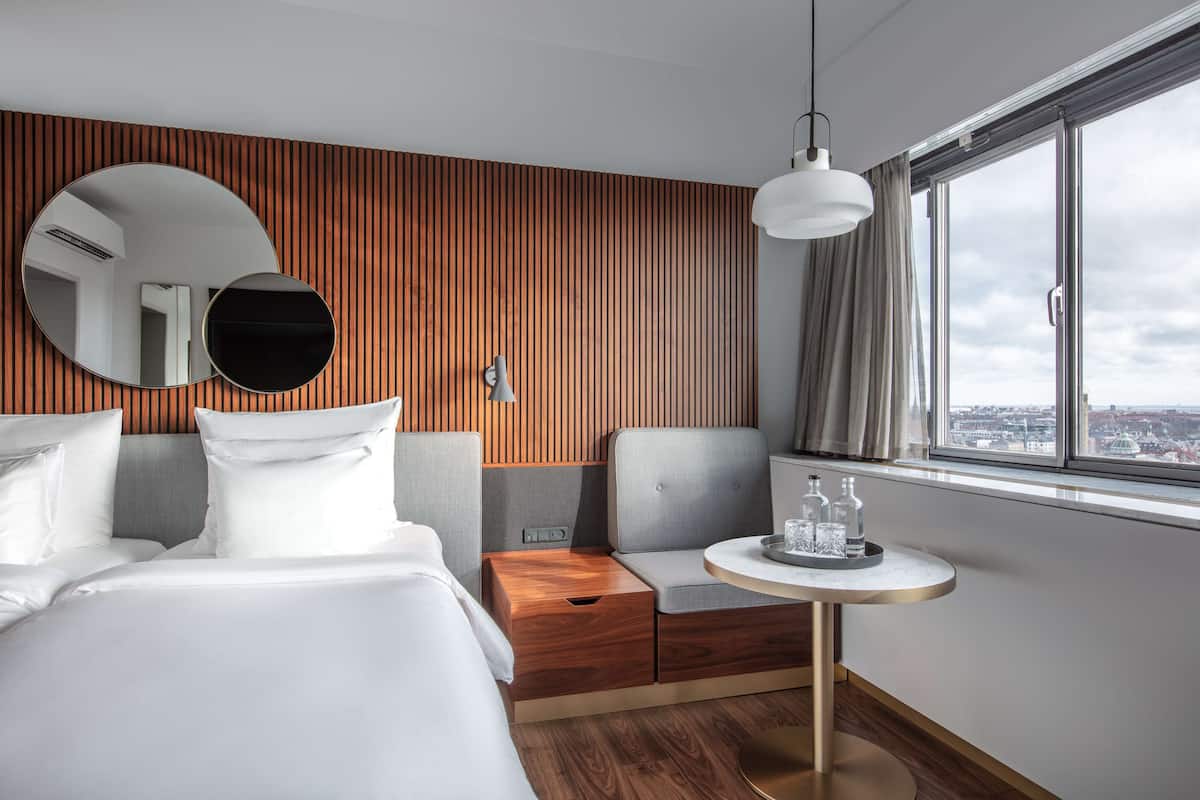 A modern hotel room with a large bed, a seating area with a small table, a large window with a city view, and a wooden panel wall.