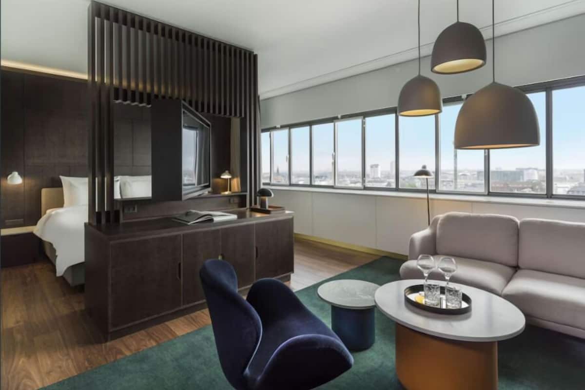 A modern hotel room with a large bed, a desk, two chairs, a sofa, and a view of the city.