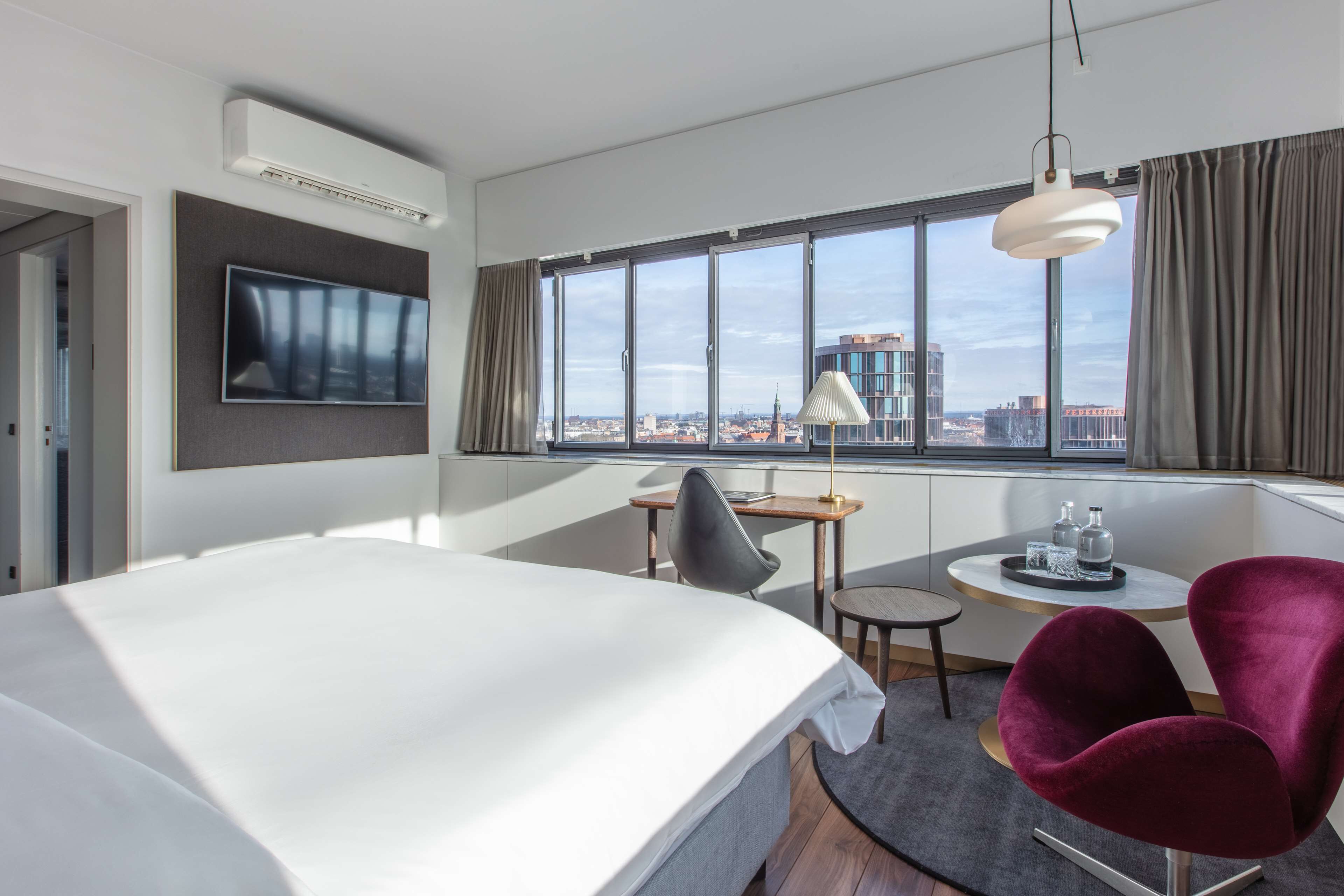 Una habitación de hotel moderna con una cama grande, un escritorio con silla, un televisor montado en la pared y un ventanal con vistas a la ciudad.