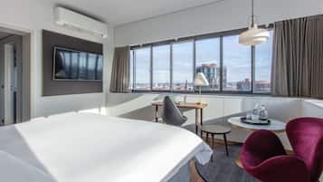 Una habitación de hotel moderna con una cama grande, un escritorio con silla, un televisor montado en la pared y un ventanal con vistas a la ciudad.