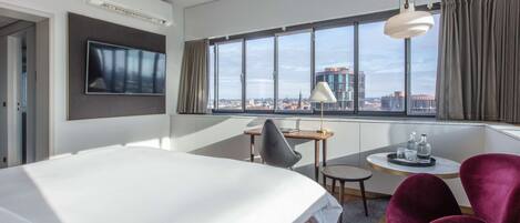 A modern hotel room with a large bed, a desk with a chair, a TV mounted on the wall, and a large window offering a city view.