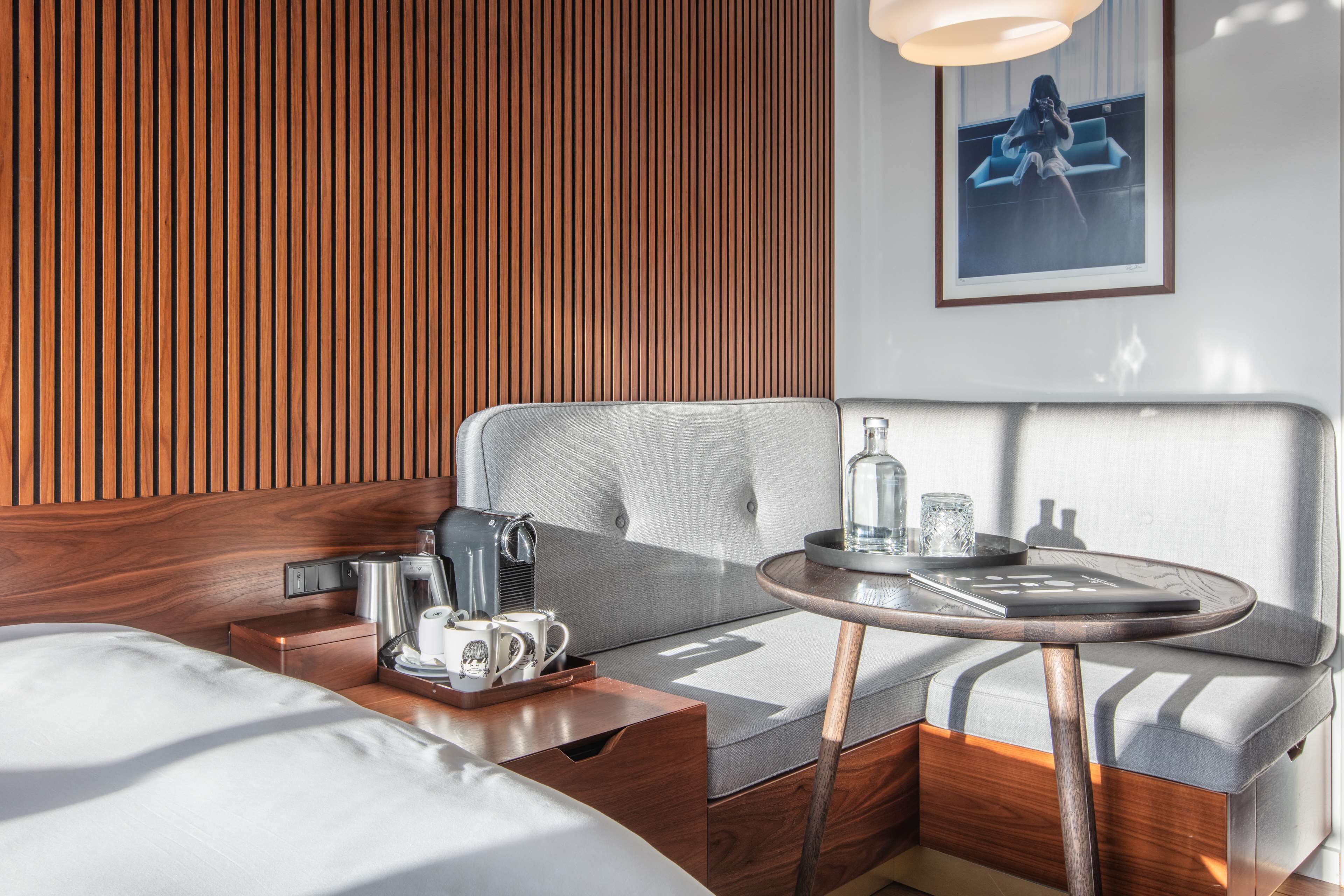 A modern hotel room with a wooden panel wall, a grey upholstered sofa, a small round table with a tray, and a coffee machine.