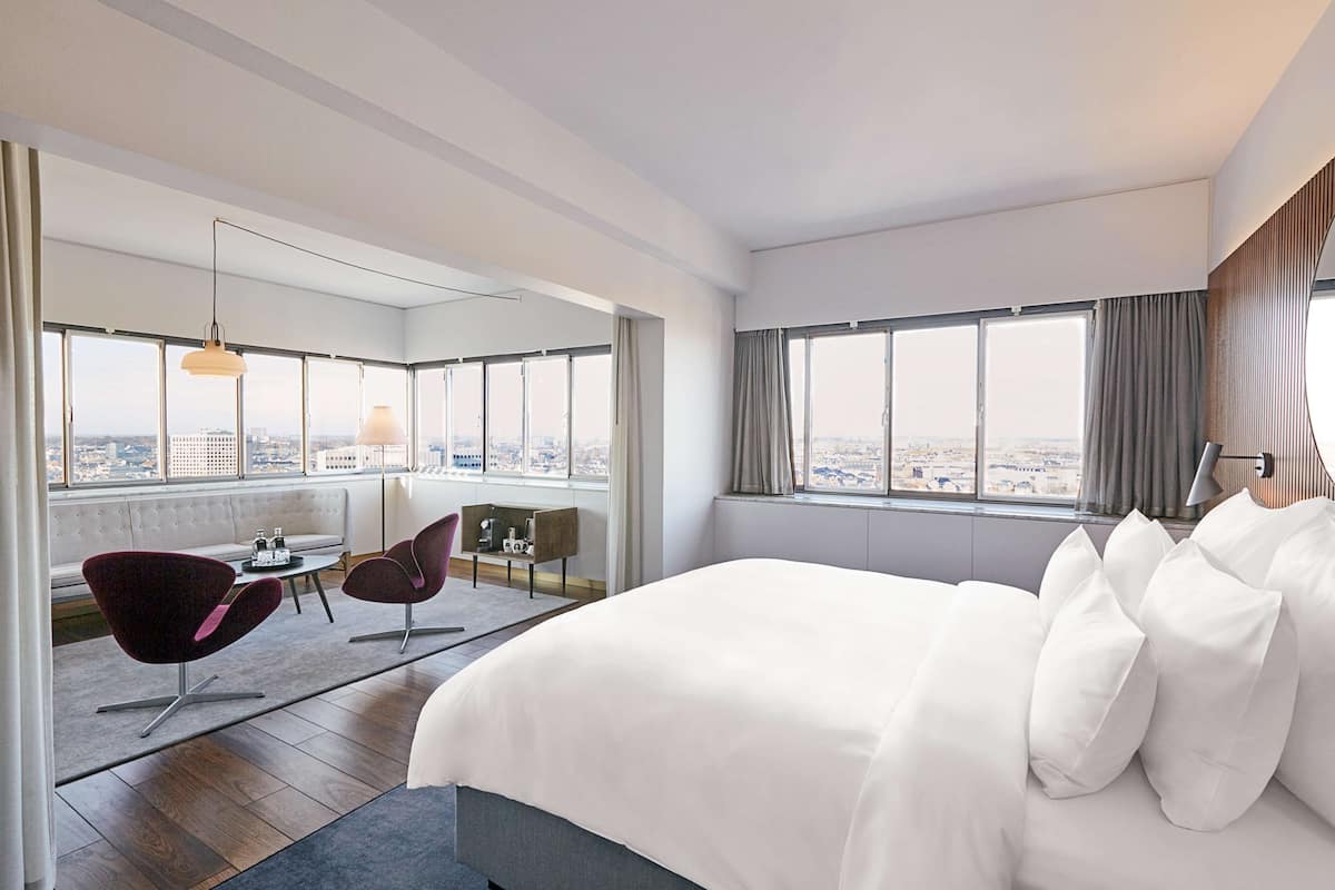 A modern hotel room with a large bed, two armchairs, a small table, and a view of the city through large windows.