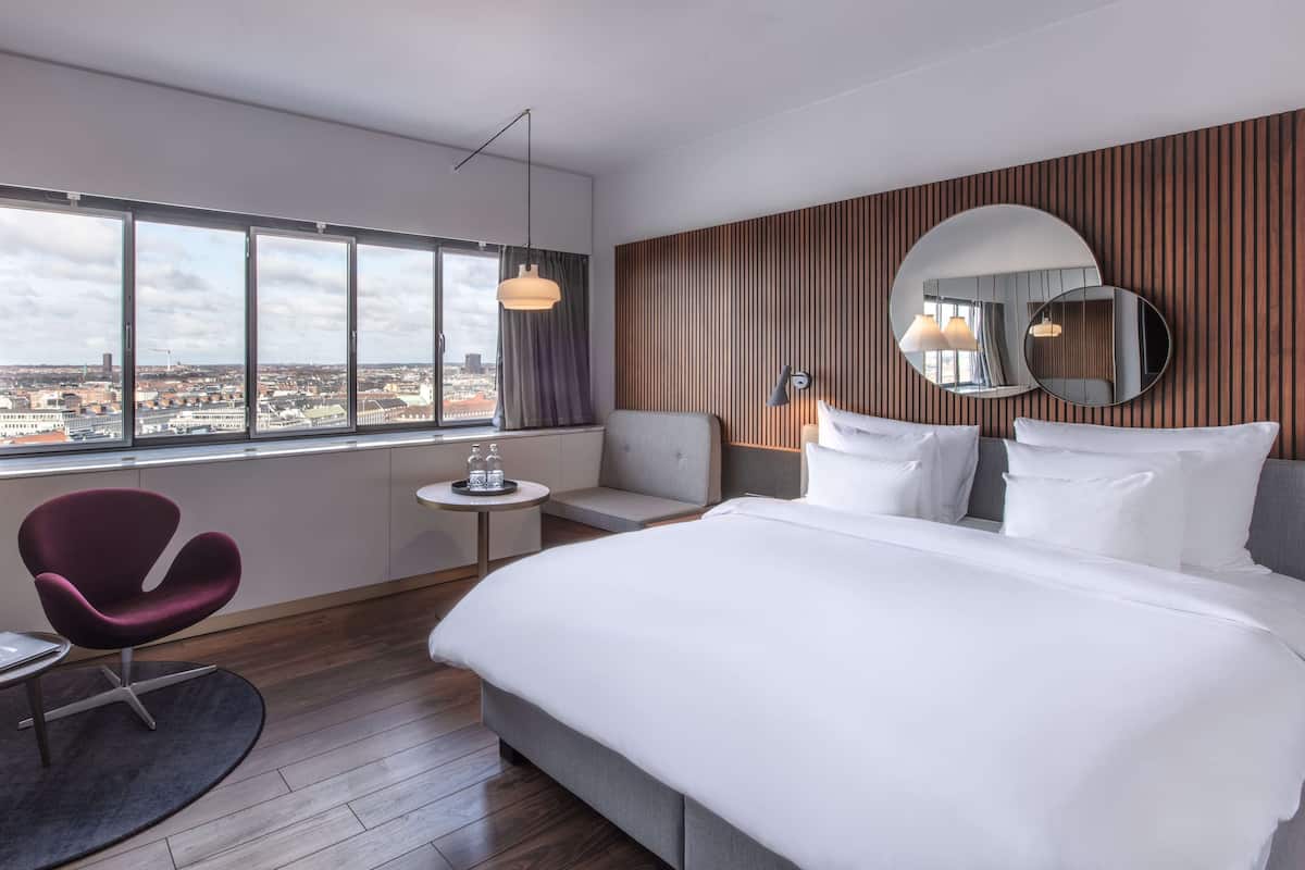 A modern hotel room with a large bed, a seating area with a chair, a small table, and a large window offering a city view.