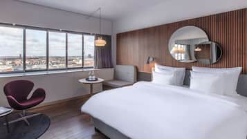 A modern hotel room with a large bed, a seating area with a chair, a small table, and a large window offering a city view.