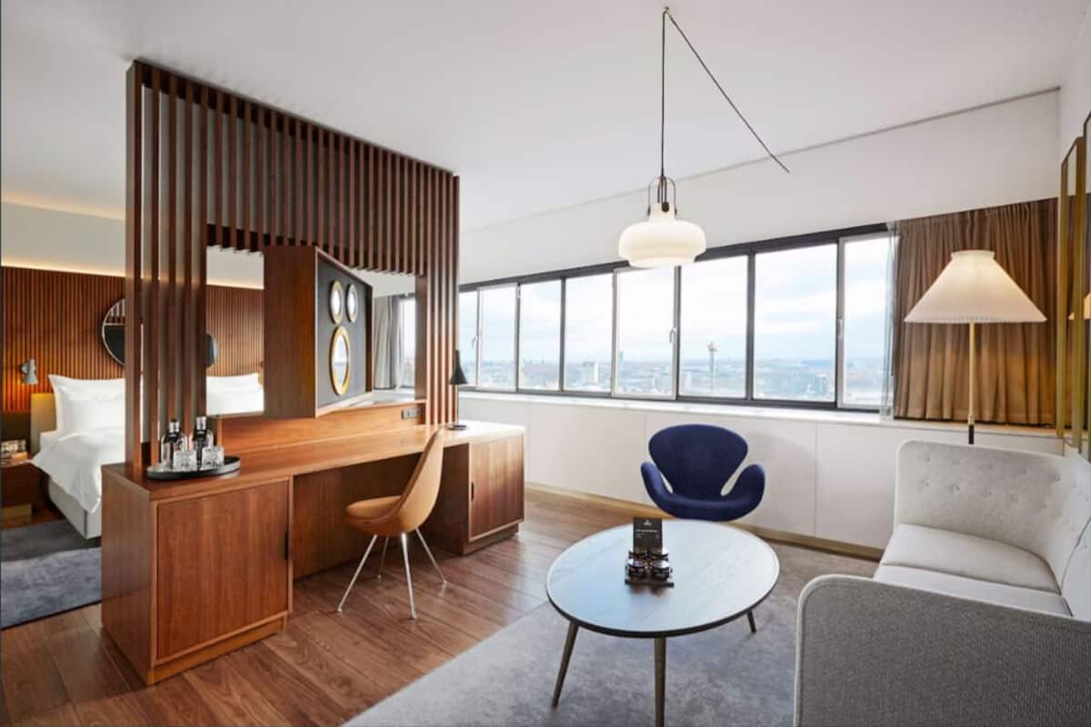 A modern hotel room with a wooden desk, a round table, a sofa, and a large window offering a city view.