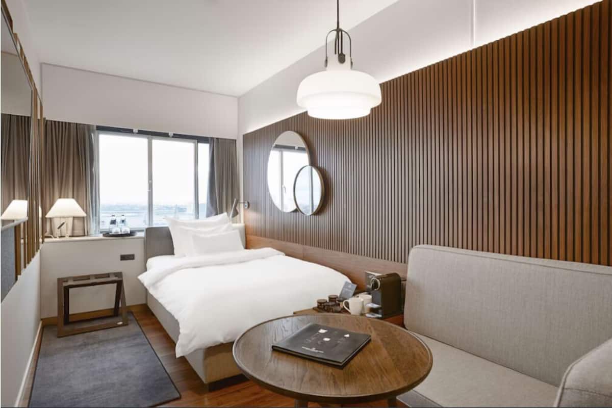A modern hotel room with a large bed, a round table, a sofa, a desk with a lamp, and a large window.