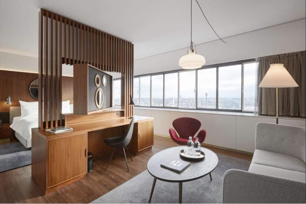 A modern hotel room with a wooden desk, a round table, a red chair, and a large window with a city view.