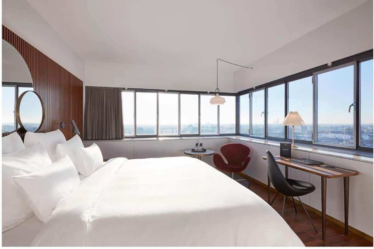 A modern hotel room with a large bed, a desk, a chair, a round mirror, and a view of the city through large windows.