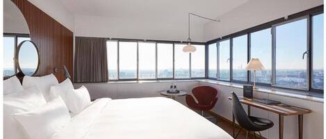 A modern hotel room with a large bed, a desk, a chair, a round mirror, and a view of the city through large windows.