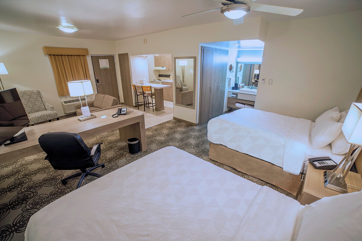 A hotel room with two beds, a desk with a chair, a small kitchenette, and a bathroom with a mirror.