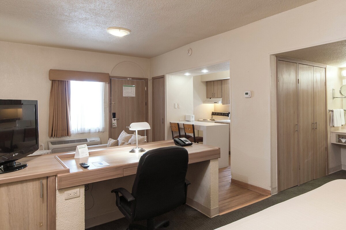 A hotel room with a desk, chair, bed, TV, and a bathroom with a sink and mirror.