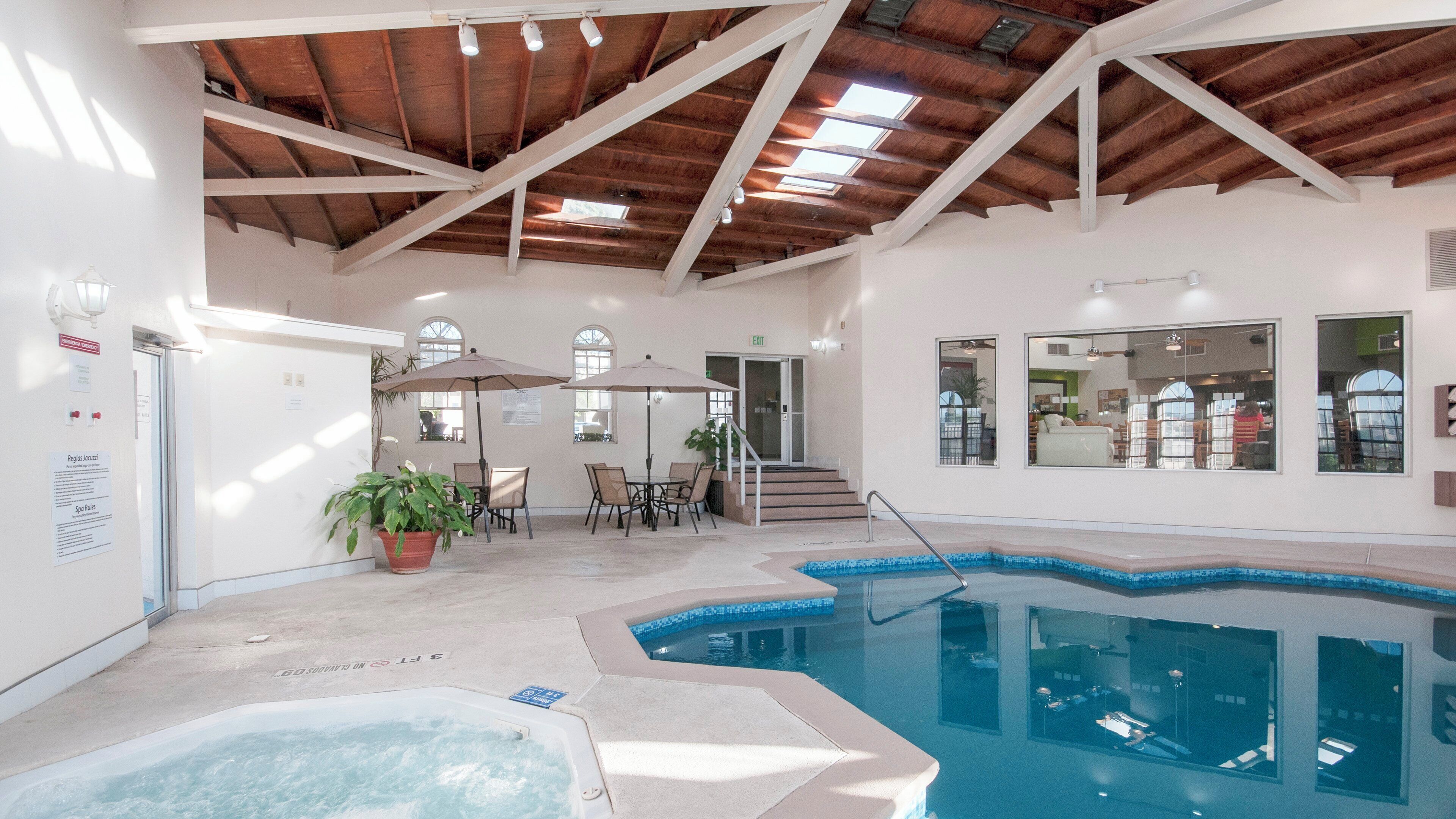 An indoor swimming pool with a hot tub, surrounded by a seating area with chairs and tables, and a staircase leading to an upper level.