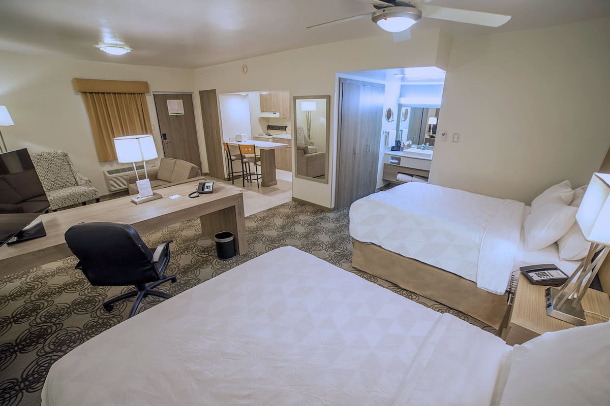 A hotel room with two beds, a desk with a chair, a small kitchenette, and a bathroom with a mirror.