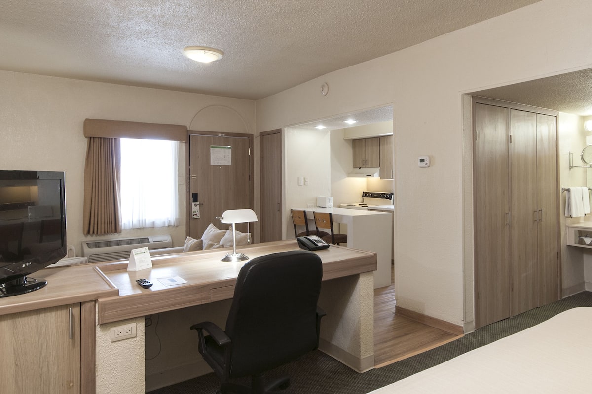 A hotel room with a desk, chair, bed, and a bathroom with a sink and mirror.