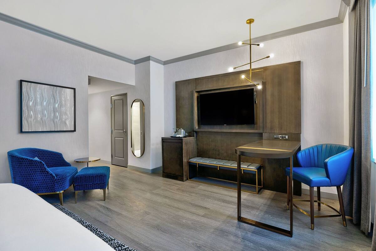 A modern hotel room with a large bed, two blue armchairs, a small table, a TV mounted on the wall, and a wooden bench.