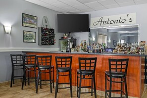 A bar area with a wooden counter, four black bar stools, a television mounted on the wall, and a sign that reads "Antonio's".