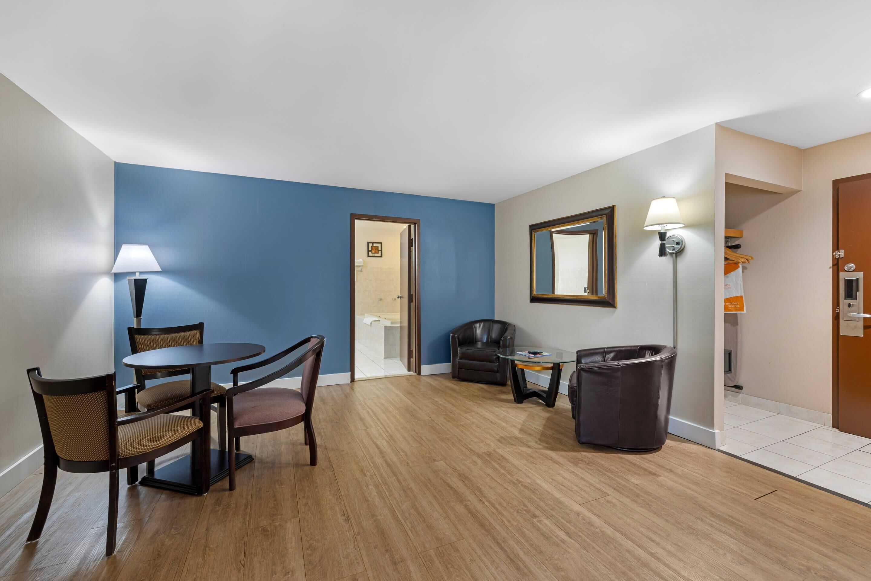 Una habitación de hotel con zona de comedor, un cubo de basura, un espejo y una puerta que da a otra habitación.