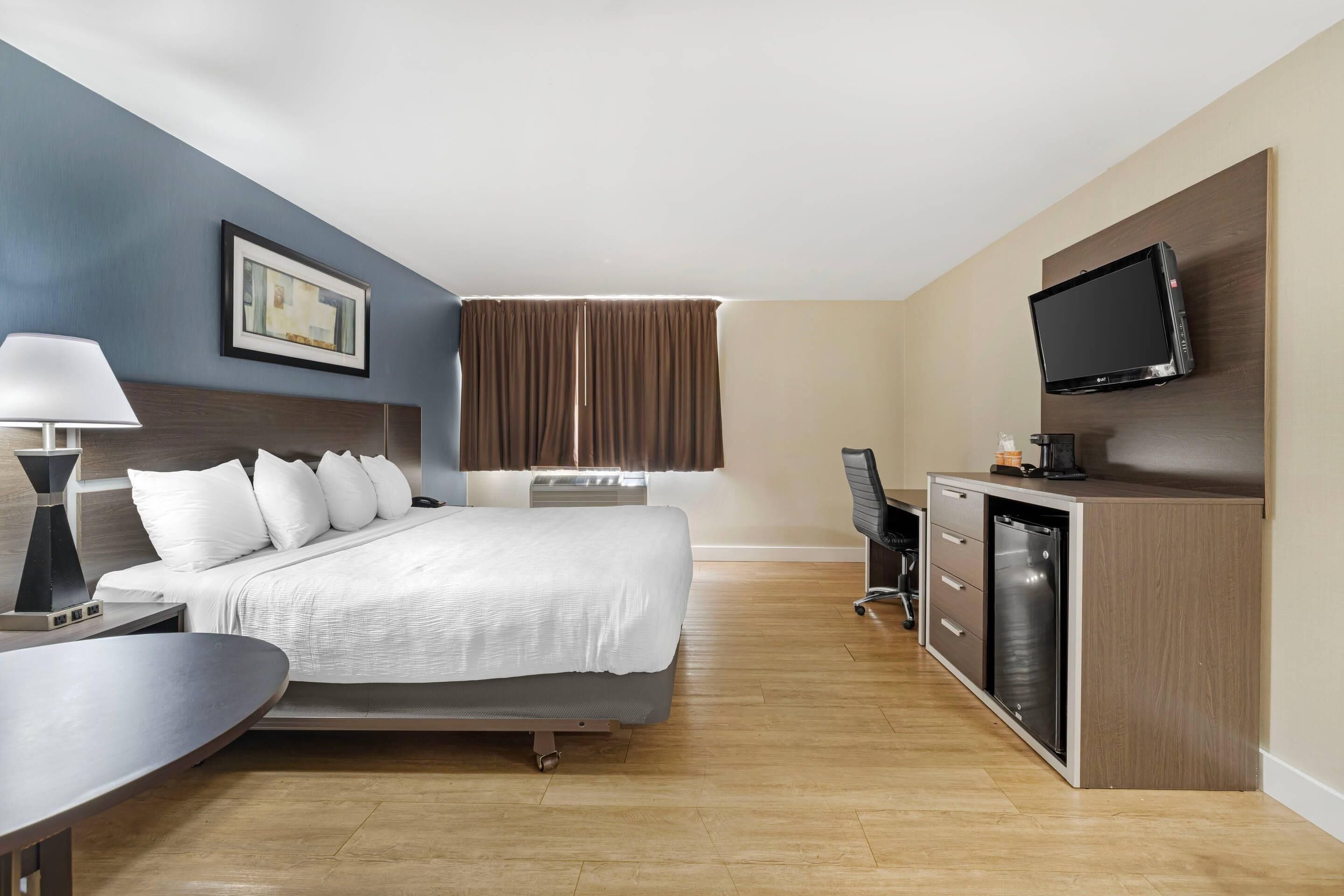 Et hotelværelse med seng, natbord, lampe, fjernsyn, skrivebord, stol og minibar.