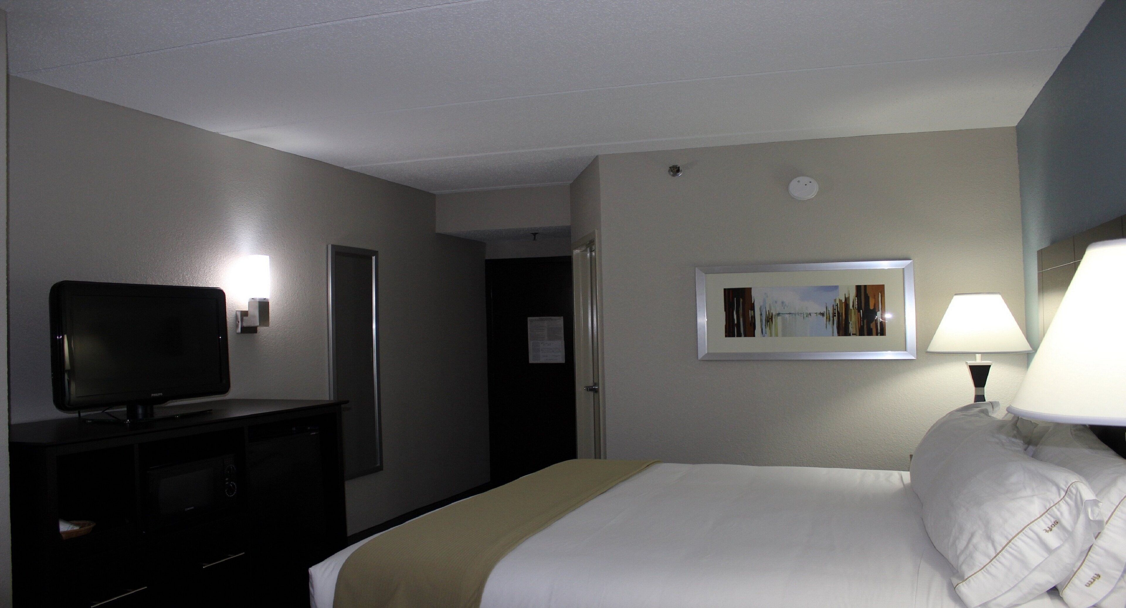 A hotel room with a bed, a television on a stand, a lamp, and a framed picture on the wall.