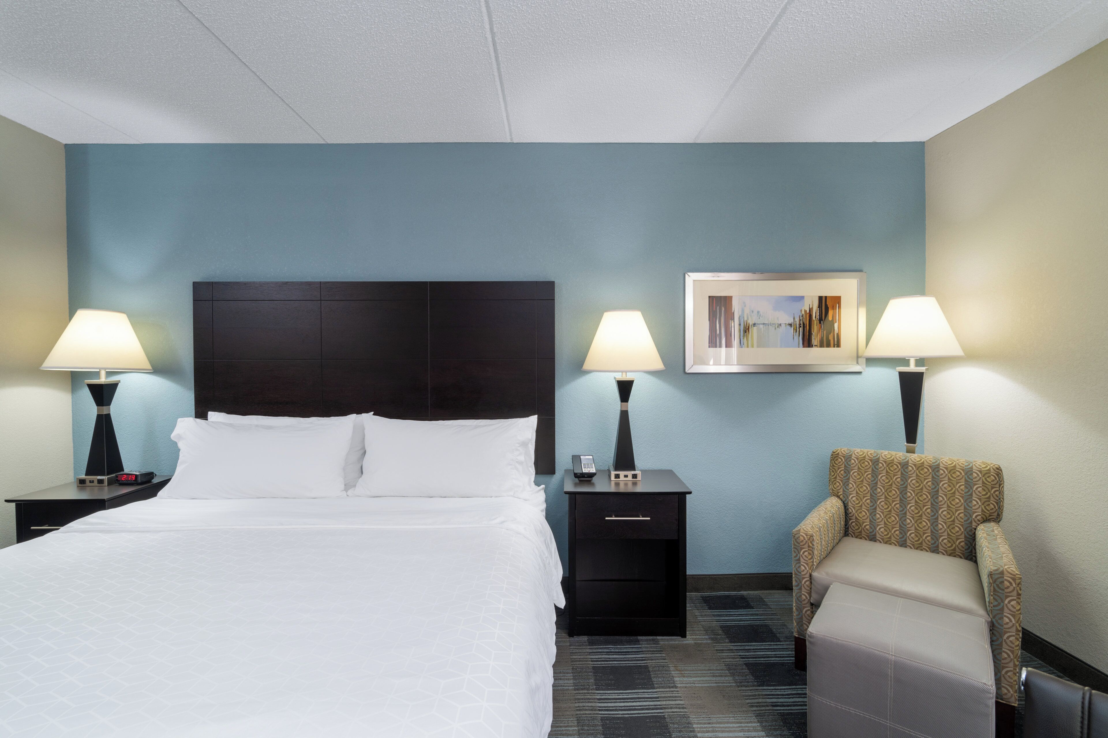 A hotel room with a bed, two bedside tables with lamps, a chair, a nightstand with a phone, and a painting on the wall.