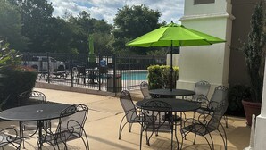 Une terrasse extérieure avec des tables et des chaises en métal, un parasol vert et une piscine en arrière-plan.