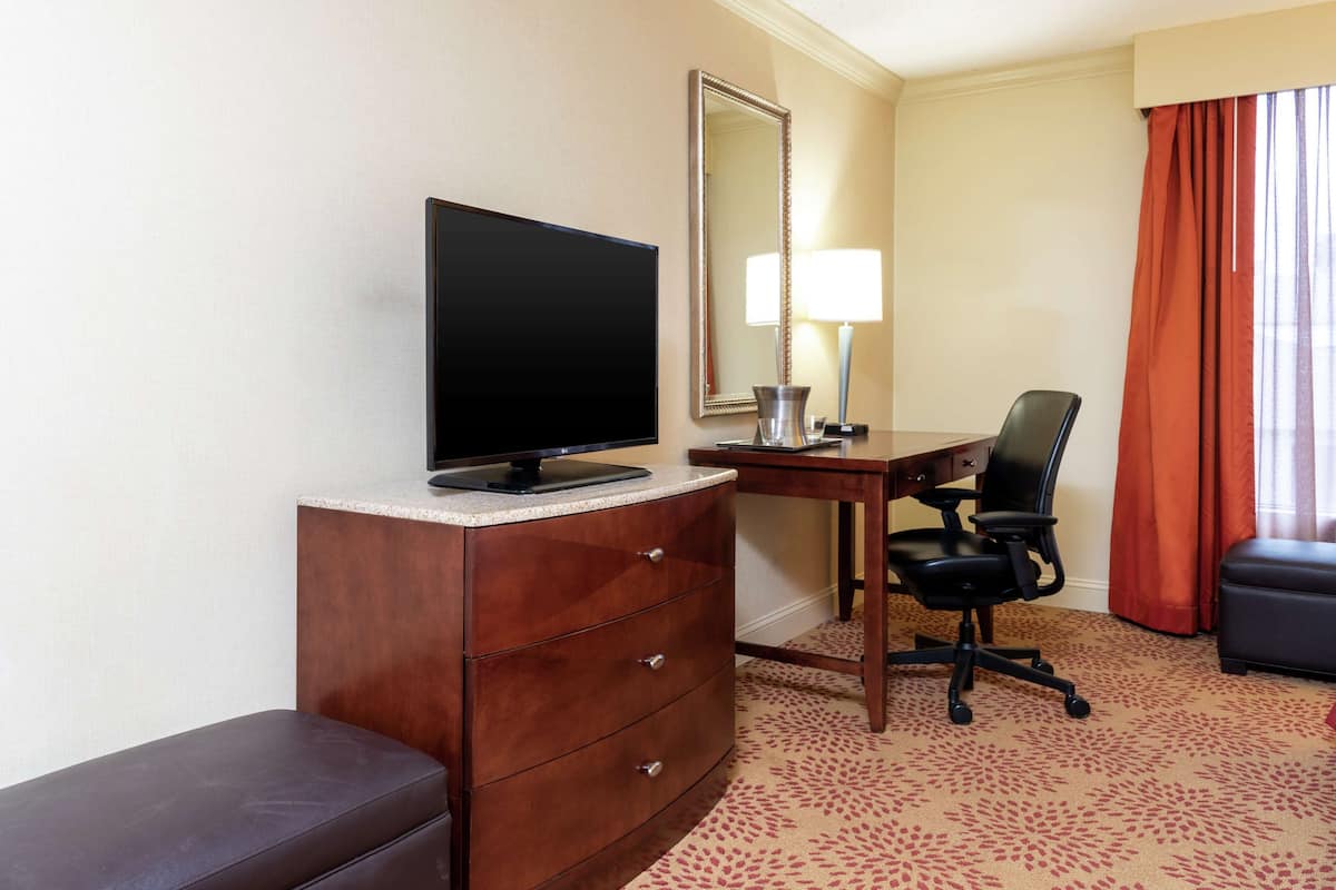 A hotel room with a TV on a dresser, a desk with a chair, a mirror, and a window with curtains.